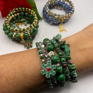 May include: Several beaded bracelets in shades of green and blue. The bracelets feature various bead sizes and textures, with gold accents and snowflake charms. One bracelet is on a wrist.