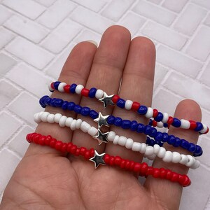 May include: Four beaded bracelets in red, white, and blue, each with a silver star charm. The bracelets are made of small, round beads and are displayed against a neutral background. The bracelets are a patriotic theme.