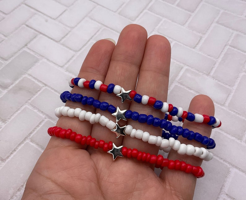 May include: Four beaded bracelets in red, white, and blue, each with a silver star charm. The bracelets are made of small, round beads and are displayed against a neutral background. The bracelets are a patriotic theme.