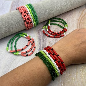 May include: Colorful beaded bracelets in shades of red, green, white, and black. Some bracelets are stacked on a gray surface, while others are worn on a wrist. The bracelets have a watermelon-like design.