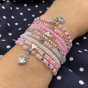 May include: A wrap bracelet featuring pink, white, and clear beads. The bracelet has silver accents and heart-shaped charms, including one with a clear stone. The bracelet is wrapped multiple times around the wrist.