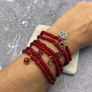 May include: A set of red beaded bracelets with silver charms. The charms are shaped like letters and have rhinestones. The bracelets are stacked on a person's wrist.