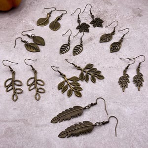 May include: A collection of bronze-toned metal earrings featuring various leaf and fern designs. Each pair is crafted with hook-style findings, showcasing intricate cut-out details and textured surfaces that mimic natural botanical shapes.