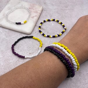 May include: Several beaded bracelets in various colors, including black, purple, white, and yellow. One bracelet is clear with black, yellow, and purple beads. The bracelets are displayed on a light gray surface.