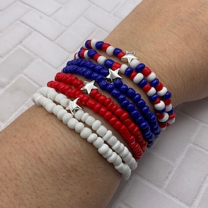 May include: A collection of beaded bracelets in red, white, and blue, with silver star charms. The bracelets are made of small, round beads and are stacked on top of each other. The color scheme evokes patriotic themes.