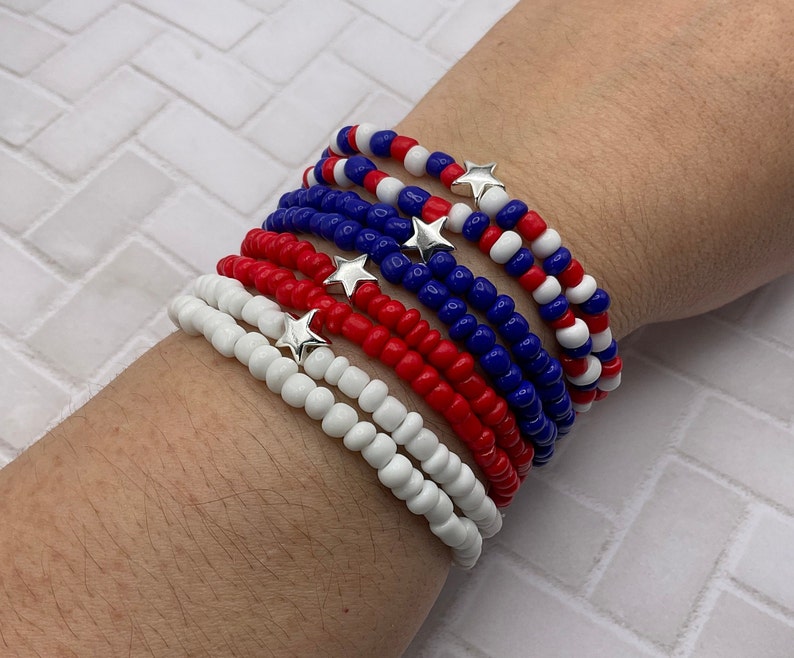 May include: A collection of beaded bracelets in red, white, and blue, with silver star charms. The bracelets are made of small, round beads and are stacked on top of each other. The color scheme evokes patriotic themes.
