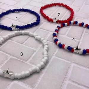 May include: Four beaded bracelets in red, white, and blue. Each bracelet features small beads and a silver star charm. The bracelets are in shades of blue, red, and white, with one bracelet combining all three colors.