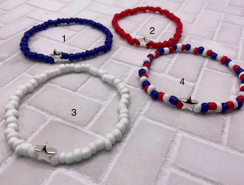 May include: Four beaded bracelets in red, white, and blue. Each bracelet features small beads and a silver star charm. The bracelets are in shades of blue, red, and white, with one bracelet combining all three colors.
