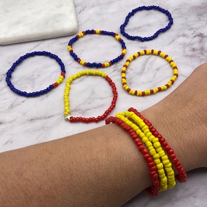 May include: A collection of beaded bracelets in various colors. The bracelets feature red, yellow, and blue beads. One bracelet has a gold star charm. Several bracelets are stacked on a wrist.