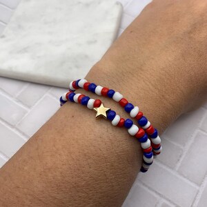 May include: Two beaded bracelets in red, white, and blue, with a gold star charm. The bracelets are worn together, showcasing a patriotic color scheme. The beads are small and round, creating a layered effect.
