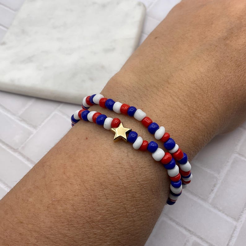 May include: Two beaded bracelets in red, white, and blue, with a gold star charm. The bracelets are worn together, showcasing a patriotic color scheme. The beads are small and round, creating a layered effect.