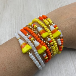 May include: A set of colorful beaded bracelets with candy corn charms. The bracelets are made with white, yellow, and orange beads. The candy corn charms are white, yellow, and orange.