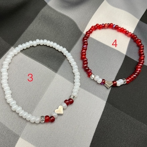 May include: Two beaded bracelets with silver heart charms. One bracelet is white with red beads and the other is red with white beads.