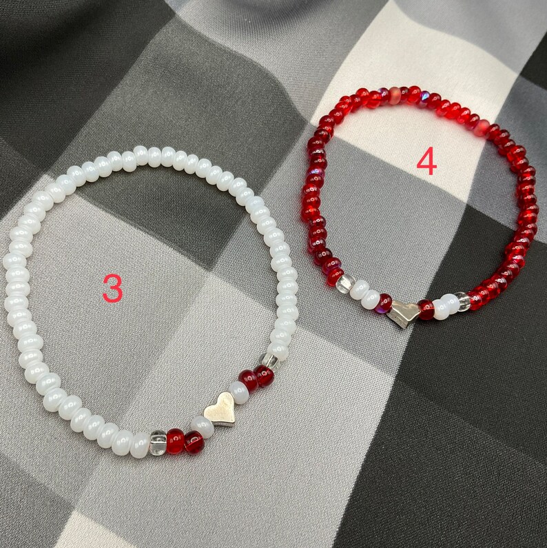 May include: Two beaded bracelets with silver heart charms. One bracelet is white with red beads and the other is red with white beads.
