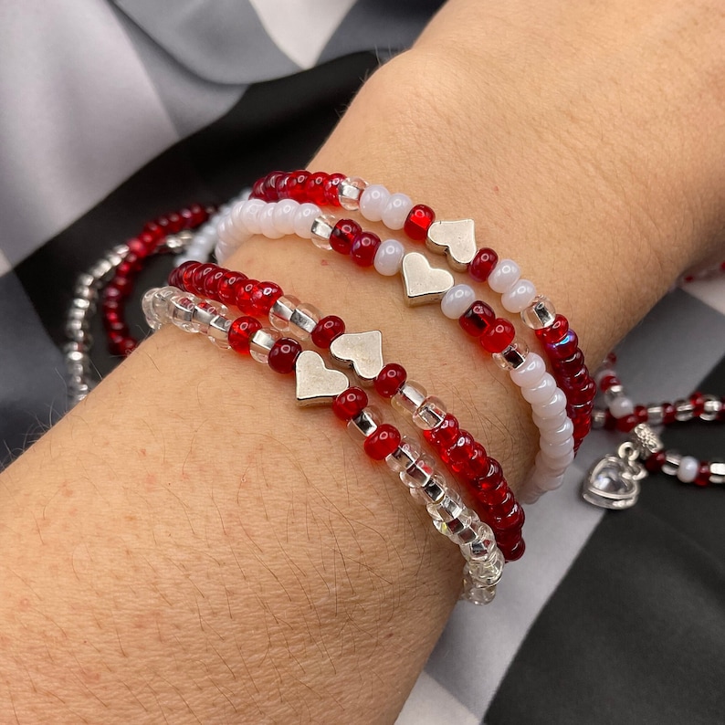 May include: A set of four bracelets with red and white beads and silver heart charms. The bracelets are stacked on a person's wrist.