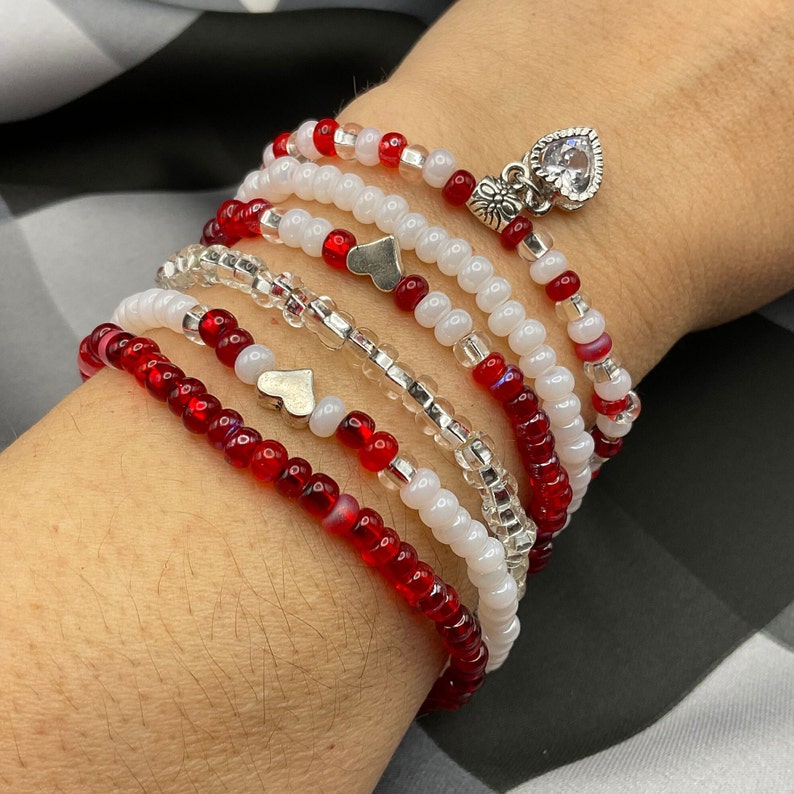 May include: A stack of red and white beaded bracelets with silver heart charms. The bracelets are layered on a person's wrist.