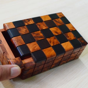 Könnte beinhalten: Eine Holz-Puzzlebox mit einem Schachbrettmuster auf der Oberseite. Die Quadrate wechseln sich in Dunkelbraun und Hellbraun ab. Die Box hat eine Schublade, die teilweise geöffnet ist und das Innere zeigt.