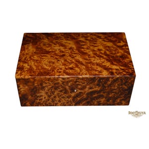 May include: A rectangular wooden box featuring a polished finish with a prominent, swirling brown and amber burl wood grain pattern. The box has a smooth, glossy surface and a small, centered white inlay detail on the front panel. Text reads ROOTTHUYA.