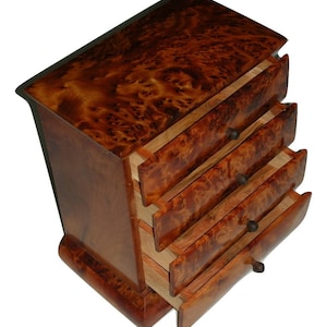 Puede incluir: Un pequeño mueble de cajones de madera con cuatro cajones, cada uno parcialmente abierto. La madera es de un color marrón oscuro intenso con un patrón de veta de raíz único. Los cajones tienen pequeños pomos oscuros. El mueble está sobre fondo blanco.