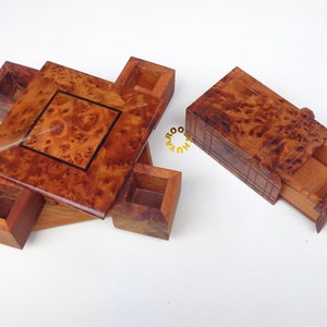May include: Wooden puzzle box with a square top and four small square compartments. The box has a natural wood grain finish with a dark square inlay. A separate rectangular box with a drawer is also visible.