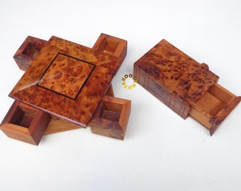May include: Wooden puzzle box with a square top and four small square compartments. The box has a natural wood grain finish with a dark square inlay. A separate rectangular box with a drawer is also visible.