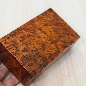 Puede incluir: Una caja de madera con un diseño geométrico intrincado. La caja está hecha de madera marrón claro con acentos marrón oscuro. La caja está cerrada y parece estar hecha de muchas pequeñas piezas de madera que encajan entre sí.