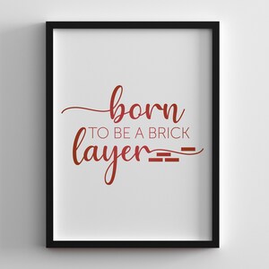 Puede incluir: Una impresión enmarcada con el texto "born to be a brick layer" en fuente cursiva roja. El texto está acentuado con un patrón de ladrillo estilizado.