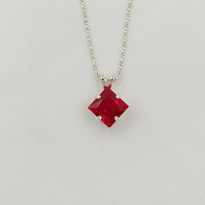 May include: A silver necklace with a square, faceted, red gemstone pendant. The pendant is a vibrant, ruby-red color and is set in a silver metal setting. The chain is a delicate, silver chain.