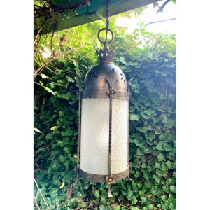 K&ouml;nnte beinhalten: Eine schwarze, h&auml;ngende Laterne aus Metall mit einem mattierten Glasschirm und dekorativen floralen Akzenten. Die Laterne h&auml;ngt an einer Kette und ist von gr&uuml;nem Laub umgeben.