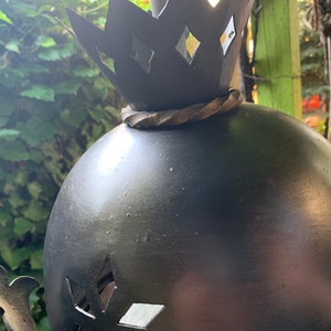 K&ouml;nnte beinhalten: Eine schwarze Metallsculptur einer Krone auf einer Kugel mit ausgeschnittenen Diamantenformen. Die Kugel steht auf einem Metallsockel.
