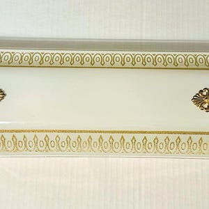 May include: A rectangular, off-white glass light fixture cover featuring a decorative gold border pattern and two ornate, oval-shaped gold metal finials centered along the length of the piece. The glass has a smooth, opaque finish.