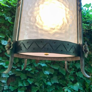 K&ouml;nnte beinhalten: Eine schwarze Metall-Laterne mit einem mattierten Glasschirm und dekorativen floralen Akzenten. Die Laterne hat ein geometrisches Design am unteren Rand.