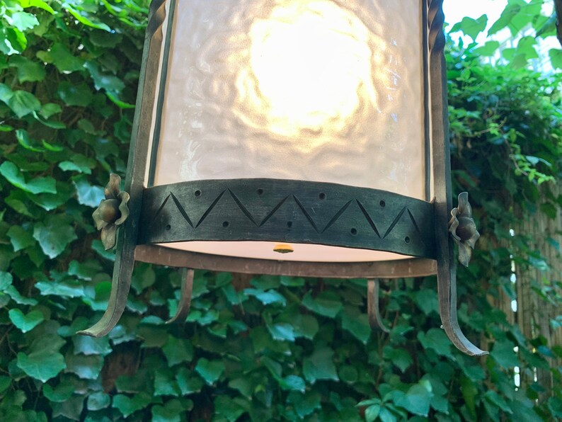 K&ouml;nnte beinhalten: Eine schwarze Metall-Laterne mit einem mattierten Glasschirm und dekorativen floralen Akzenten. Die Laterne hat ein geometrisches Design am unteren Rand.