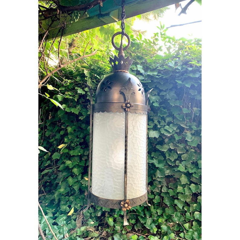 K&ouml;nnte beinhalten: Eine schwarze, h&auml;ngende Laterne aus Metall mit einem mattierten Glasschirm und dekorativen floralen Akzenten. Die Laterne h&auml;ngt an einer Kette und ist von gr&uuml;nem Laub umgeben.