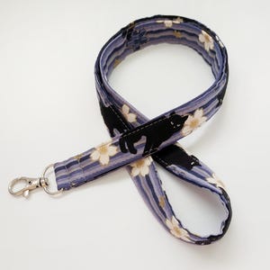 May include: A fabric lanyard with a silver-colored clasp. The lanyard features a pattern of black cats and white flowers on a striped background of blue, purple, and gray.