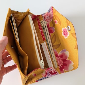 May include: A mustard yellow fabric wallet with a floral pattern featuring pink and purple blossoms. The interior includes multiple card slots, a zippered compartment, and open pockets containing paper currency and various cards.