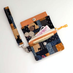 May include: A fabric wristlet wallet featuring a patchwork design of black cats, floral patterns, and geometric shapes in shades of navy, tan, and orange. The wallet includes a zippered compartment, a snap closure, and a detachable wrist strap.
