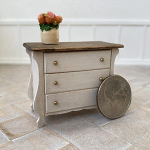May include: A miniature three-drawer chest in a light wood finish with brass knobs. The top is stained a darker brown. A small vase with pink and orange flowers sits on top. A coin is next to the chest for scale.