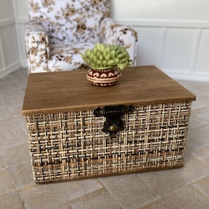 以下が含まれることがあります： 木製の天板と装飾的な留め金が付いた、ミニチュアの織り収納チェスト。緑色の多肉植物が入った、赤と白の模様の小さな丸い鉢が上に置かれています。チェストは織られた天然繊維で作られています。