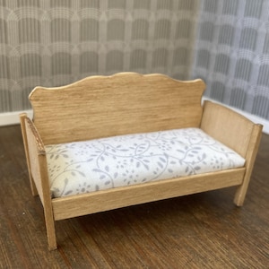 May include: A miniature wooden sofa with a light-colored finish. The seat cushion features a white fabric with a gray floral pattern. The sofa has a curved back and armrests, designed for a dollhouse or miniature setting.