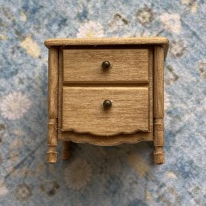 May include: A small wooden nightstand with two drawers and simple metal knobs. The nightstand is a light brown color and has a slightly distressed finish.
