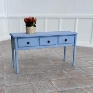 May include: A miniature blue painted wooden console table with three drawers and black knobs. A small vase with pink and brown flowers sits on top. The table has a scalloped edge and four legs.
