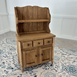 Op de afbeelding: Een miniatuur houten buffetkast met een medium bruine afwerking, een hoog achterpaneel met een open plank, drie kleine lades aan de bovenzijde en twee onderste kastdeurtjes voorzien van kleine, ronde knoppen als handgrepen.