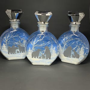May include: Three round glass bottles with silver diamond-shaped stoppers, each containing a string of fairy lights. The front of each bottle features a winter scene with silhouettes of bears in a snowy forest with white trees and blue sky.