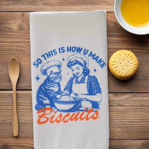 May include: A white kitchen towel with a blue and orange design. The design features a woman and a cat wearing chef hats, making biscuits. The text on the towel reads "So this is how u make biscuits".