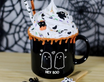 May include: A black Halloween-themed mug with white whipped cream, sprinkles, and decorative accents. The mug features two ghost illustrations and the text "HEY BOO". A small wooden spoon and a plastic spider are also present.