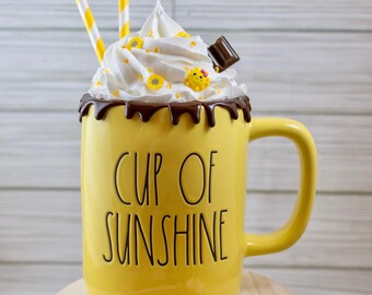 May include: A yellow mug with the words "CUP OF SUNSHINE" in black lettering, topped with whipped cream, chocolate drizzle, and decorative accents. Two yellow and white striped straws are in the mug. A wooden spoon with whipped cream sits on a small wooden stool.