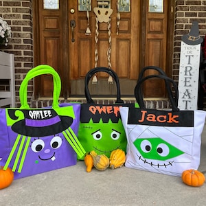May include: Three Halloween trick-or-treat bags with personalized names: Oakley, Owen, and Jack. The bags are decorated with different Halloween characters: a witch, a Frankenstein monster, and a mummy. The bags are sitting on a concrete porch with a pumpkin in front of each bag.