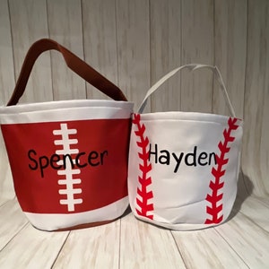 May include: Two fabric baskets, one designed as a football and the other as a baseball. The football basket is red with white stripes and the name "Spencer". The baseball basket is white with red stitching and the name "Hayden". Both have handles.