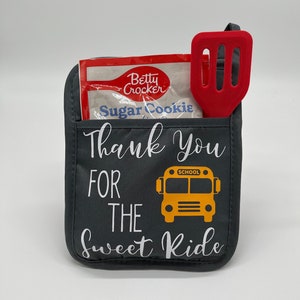 Puede incluir: Manopla de horno gris con el texto "Thank You For The Sweet Ride" y un gr&aacute;fico de autob&uacute;s escolar amarillo. Una esp&aacute;tula roja y un paquete de mezcla para galletas Betty Crocker Sugar Cookie est&aacute;n en el bolsillo de la manopla de horno.